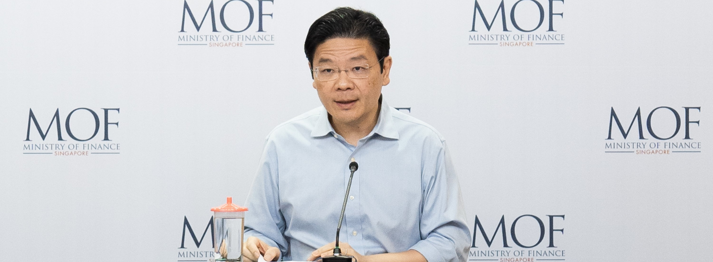 Asian man in glasses speaking at a podium; MOF Ministry of Finance Singapore logos in background.